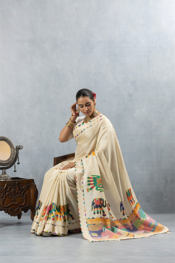 Model seated gracefully in the Vanya Sarees off-white cotton silk saree, with the colorful elephant print border beautifully displayed.