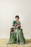 Model seated gracefully in the Vanya Sarees green Banarasi saree, holding a lotus flower to complement the blouse embroidery.